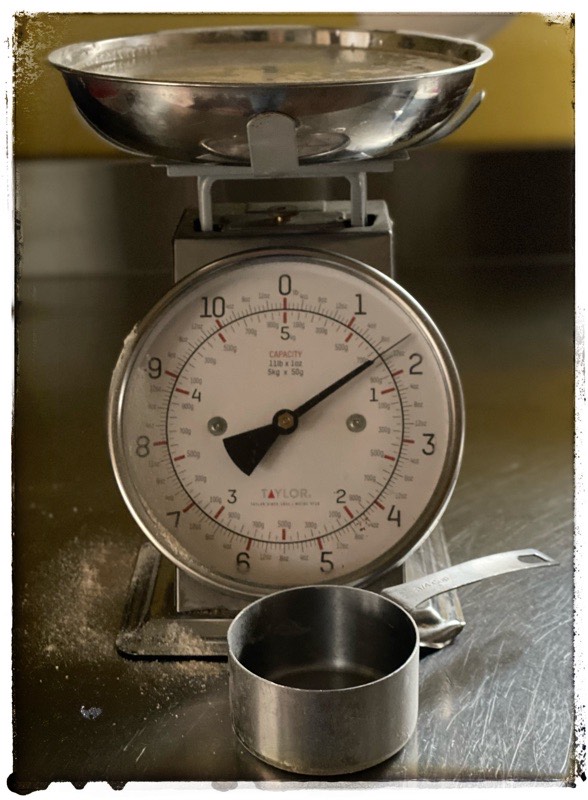 Manual scales. Showing 780g of water in metal bowl. Water has yeast floating on top. Empty stainless steel I cup measurement sits at the base of the scales. Scales are standing on stainless steel counter. 