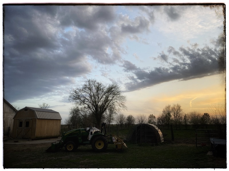 Late evening with feathery windy dark clouds. JLow light. ohn Deere tractor in the mid foreground. Feed hut to the left. Metal hoop house to the right. Hints of pinks and pale blue in the sky. Trees lining the horizon. 