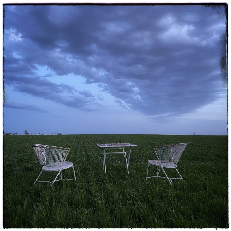 Field of wheat in the early evening looking South. Grey clouds gathering overhead. Two wrought iron chairs and a table in the field. 