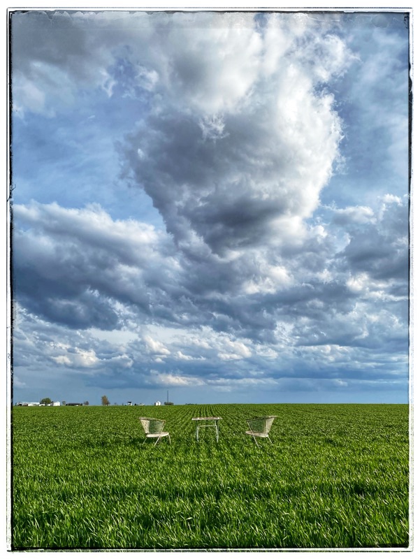 Midwest sky. Midwest clouds. Flat country. Wheat fields with two chairs and a table in the foreground. Grey and white clouds above. 